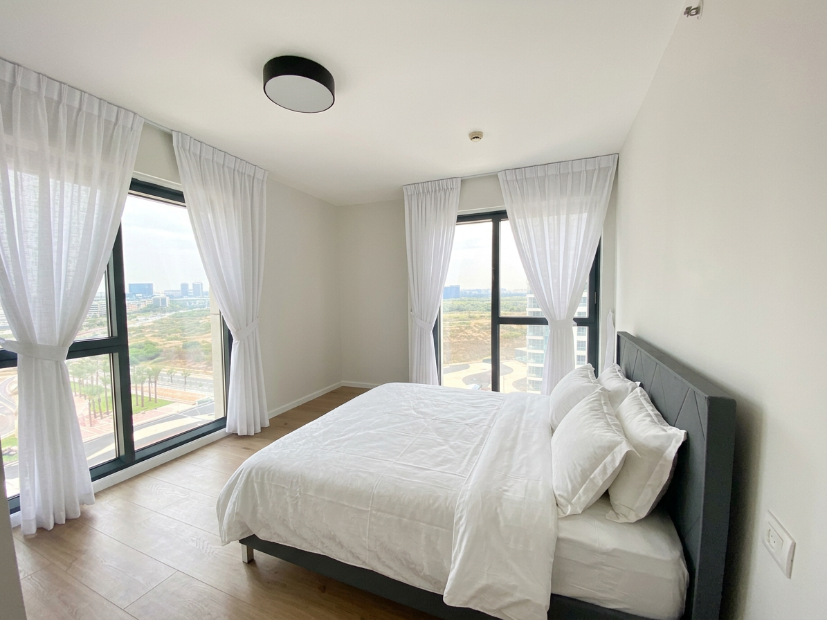 Bright corner bedroom with white curtains, grey upholstered bed and a city view window